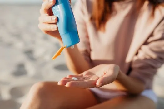 Korean adult applying sunscreen to forearm before summer outdoor activity, hat and water bottle nearby, bright realistic photo, no brand logos