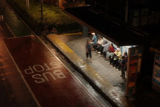Korean commuter at shaded bus stop drinking water during heatwave, checking condition before boarding, realistic summer editorial photo, no readable signs