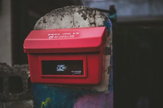 Sealed paper envelope labeled 폐의약품 beside red mailbox, no syrup bottle sign, Korean neighborhood street, realistic public service photo | 폐의약품 봉투를 우체통에 넣기 전 확인하는 장면