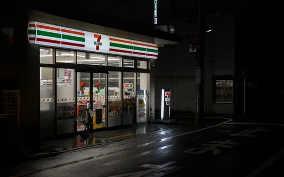 Korean adult checking official medicine list on smartphone outside a closed pharmacy and convenience store, rainy evening, realistic editorial photo, no readable logos